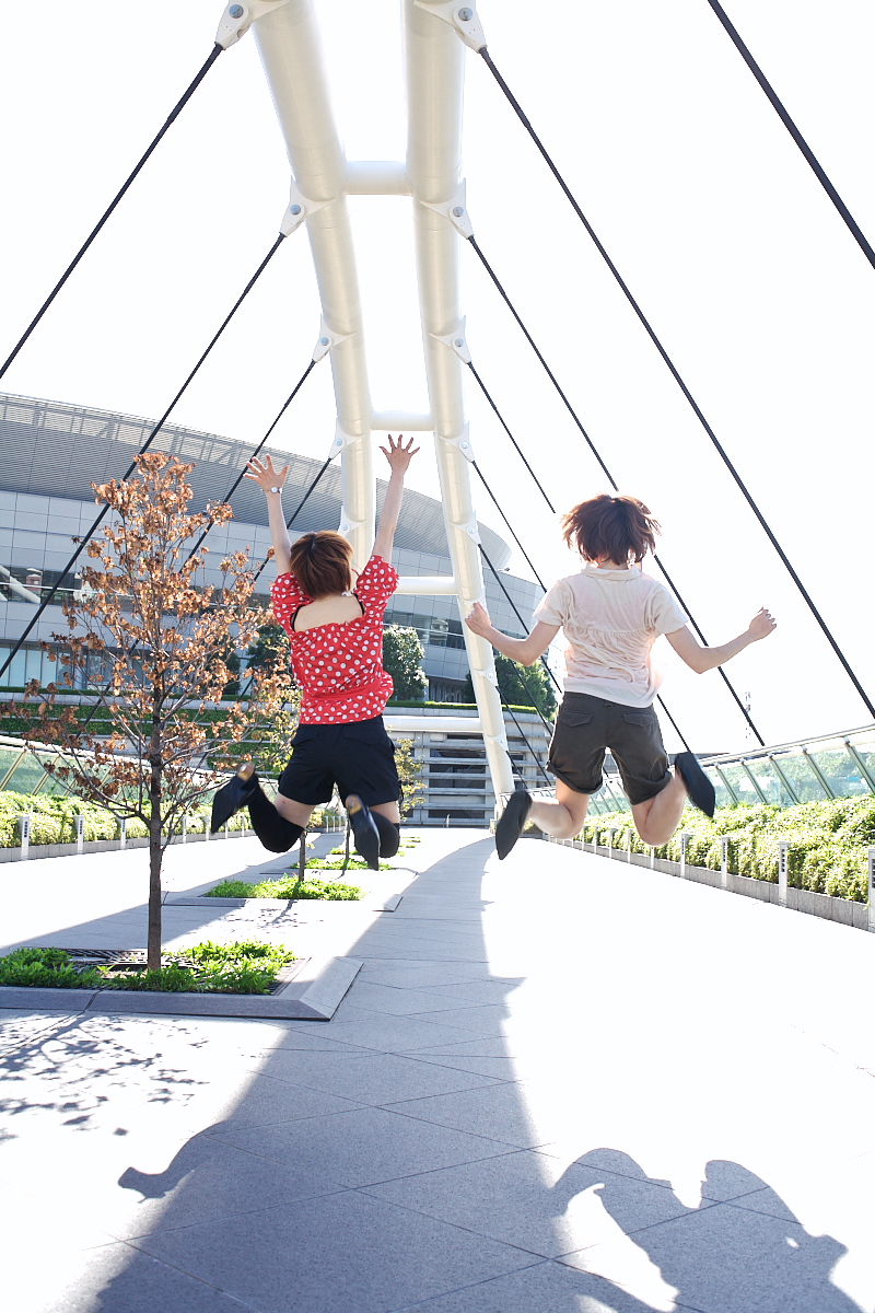 2girls ari_(model) asian black_shorts bridge brown_hair building chippi cosplay dyed_hair female_focus from_behind highres jumping multiple_girls outdoors photo_(medium) plant polka_dot polka_dot_shirt shadow shirt short_hair shorts thighhighs tree