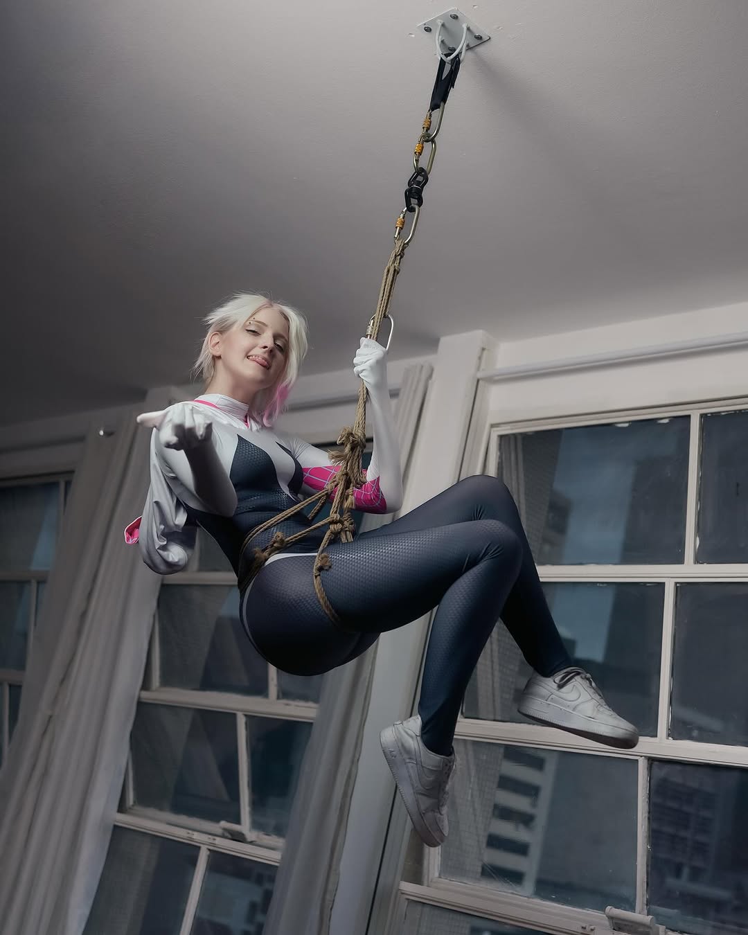 1girl arm_support ass black_bodysuit blue_eyes bodysuit brazilian_(nationality) breasts brown_hair cosplay curvy freckles gwen_stacy highres hood hooded_bodysuit indoors looking_at_viewer marvel memepantazis naughty_face photo_(medium) shibari shiny_clothes smile spider-gwen spider-gwen_(cosplay) spider-man_(series) wide_hips