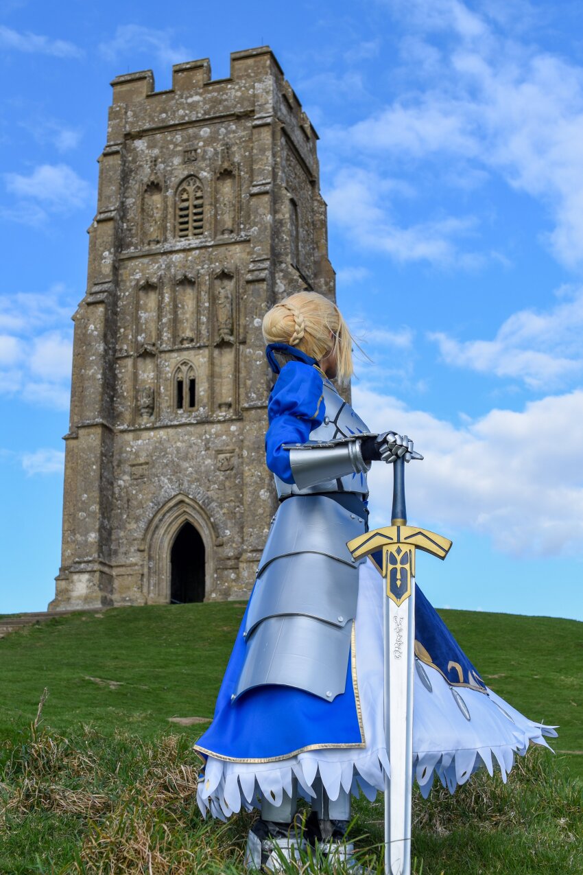 1girl absurdres armor armored_boots armored_dress artoria_pendragon_(fate) blue_dress blue_ribbon blue_sky boots breastplate bun_with_braided_base cloud cloudy_sky commentary_request cosplay_photo day dress england excalibur_(fate/stay_night) facing_away fate/stay_night fate_(series) gauntlets glastonbury_tor grasslands hair_bun hair_ribbon hand_on_hilt highres hino_yoriko juliet_sleeves long_dress long_sleeves looking_back no_ahoge on_grass outdoors photo_(medium) planted planted_sword planted_weapon puffy_sleeves real_world_location ribbon saber_(fate) sidelocks single_hair_bun sky solo standing sword tower turning_head united_kingdom updo weapon