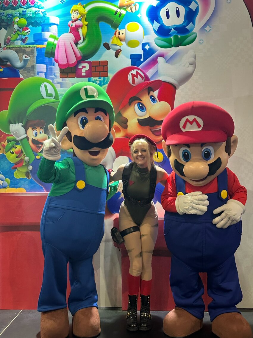 1girl 2boys arm_tattoo bare_legs bare_shoulders blonde_hair blue_eyes braid brazilian_(nationality) breasts cammy_white cammy_white_(cosplay) capcom clenched_hands cosplay curvy dress elephant_mario fingerless_gloves gloves goomba grin happy hat highleg highleg_leotard highres legs leotard long_hair looking_at_viewer luigi mario mario_(series) medium_breasts memepantazis multiple_boys nintendo pale_skin photo_(medium) pink_dress prince_florian princess_peach real_life sleeveless smile street_fighter street_fighter_6 super_mario_bros._wonder super_smash_bros. tattoo toad_(mario) twin_braids warp_pipe wide_hips wonder_flower yoshi