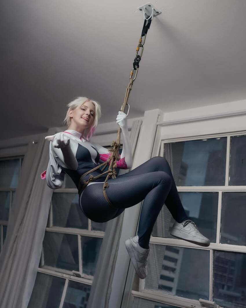 1girl arm_support ass black_bodysuit blue_eyes bodysuit brazilian_(nationality) breasts brown_hair cosplay curvy freckles gwen_stacy highres hood hooded_bodysuit indoors looking_at_viewer marvel memepantazis naughty_face photo_(medium) shibari shiny_clothes smile spider-gwen spider-gwen_(cosplay) spider-man_(series) wide_hips
