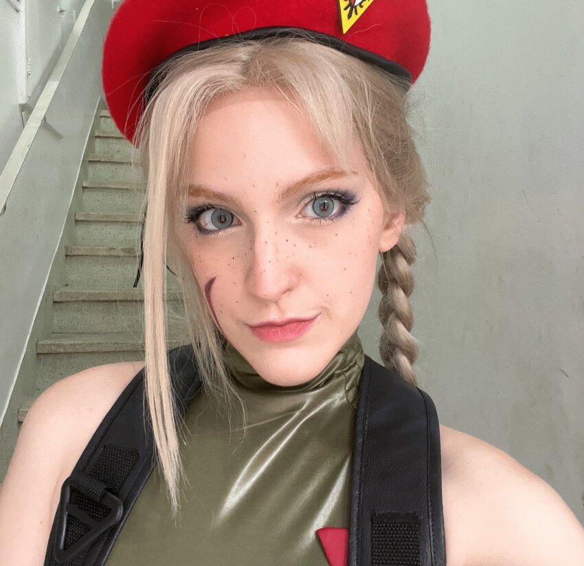 1girl :&gt; bare_shoulders blonde_hair blue_eyes braid brazilian_(nationality) breasts cammy_white cammy_white_(cosplay) cosplay freckles green_leotard hat highres leotard long_hair memepantazis pale_skin photo_(medium) real_life sleeveless smile street_fighter street_fighter_6 twin_braids twitter_username upper_body