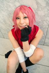 1girl ari arm_warmers bare_shoulders black_gloves black_socks cosplay eyelashes female_focus gloves green_eyes hand_up haruno_sakura haruno_sakura_(cosplay) headband headdress highres holster konohagakure_symbol lips looking_at_viewer looking_up miniskirt naruto_(series) naruto_shippuuden photo_(medium) pink_hair red_shirt shirt short_hair sitting skirt sleeveless sleeveless_shirt smile socks solo thigh_holster white_skirt