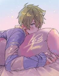 1boy blonde_hair blue_eyes blurry blush commentary fingernails hair_between_eyes hugging_object idolmaster idolmaster_side-m kashiwagi_tsubasa light_particles looking_at_viewer lying male_focus nana_tetra on_bed on_stomach pillow pillow_hug pink_pillow pink_sweater purple_sweater short_hair solo star_(symbol) sweater symbol-only_commentary two-tone_sweater upper_body