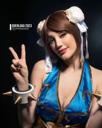 1girl alternate_costume argentine_(nationality) bare_hips bare_legs bare_shoulders blue_dress blue_eyes bracelet braid breasts brown_hair capcom chun-li chun-li_(cosplay) cleavage cosplay double_bun dress fingernails hair_bun highleg highres instagram_username jewelry lips lipstick long_hair looking_at_viewer makeup medium_breasts no_bra pelvic_curtain photo_(medium) revealing_clothes shiny_clothes short_dress simple_background smile sophie_valentine spiked_bracelet spikes standing street_fighter street_fighter_iv_(series) upper_body v