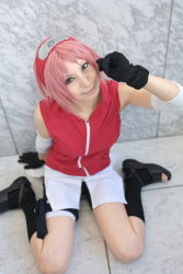 1girl ari arm_warmers bandages bare_shoulders bike_shorts black_gloves black_shoes black_socks blurry cosplay depth_of_field eyelashes female_focus gloves green_eyes hand_up haruno_sakura haruno_sakura_(cosplay) head_tilt headband headdress highres holster konohagakure_symbol lips looking_at_viewer looking_up miniskirt naruto_(series) naruto_shippuuden photo_(medium) pink_hair red_shirt shirt shoes short_hair shorts shorts_under_skirt side_slit sitting skirt sleeveless sleeveless_shirt smile socks solo thigh_holster thigh_strap toeless_legwear toes wariza white_skirt